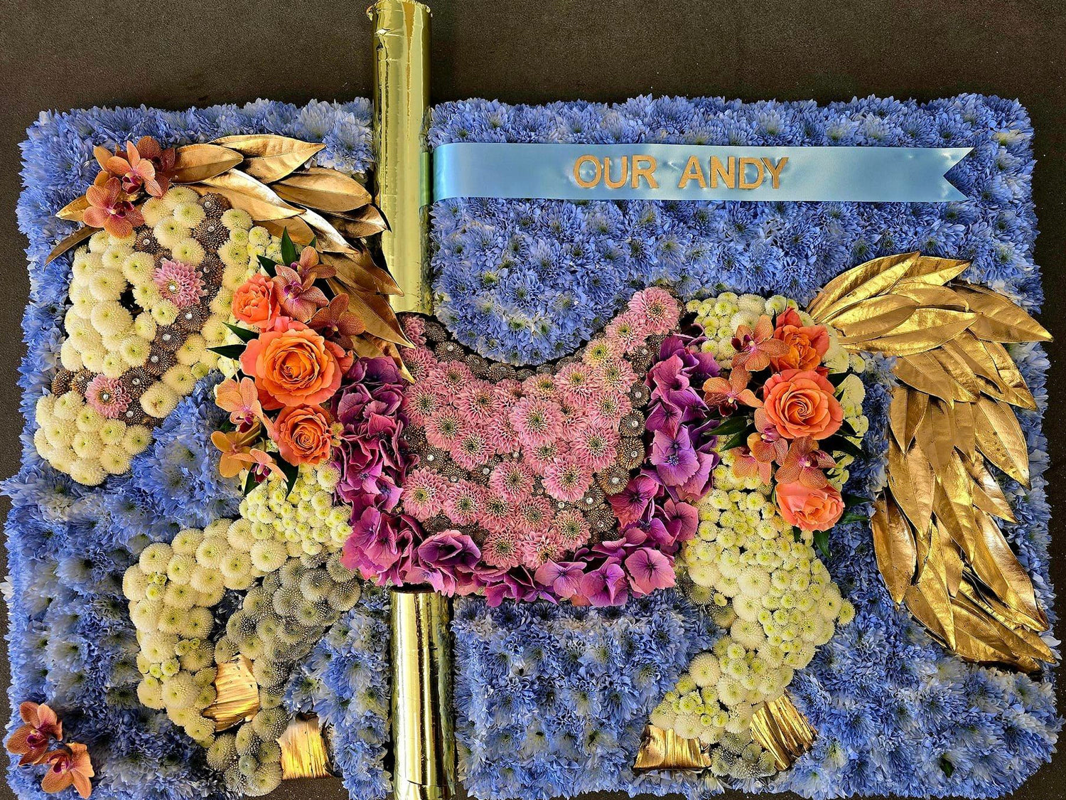 A custom floral arrangement shaped like a fairground horse, featuring blue flowers for the body, purple and pink flowers for the mane and tail, orange roses, gold leaves 