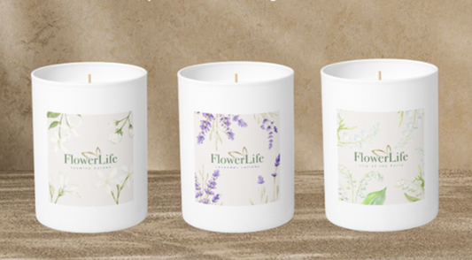 Three white soy candles in cylindrical jars with floral labels, displayed side by side on a wooden surface against a textured beige background. The labels read 'FlowerLife' and feature botanical illustrations in green and purple.