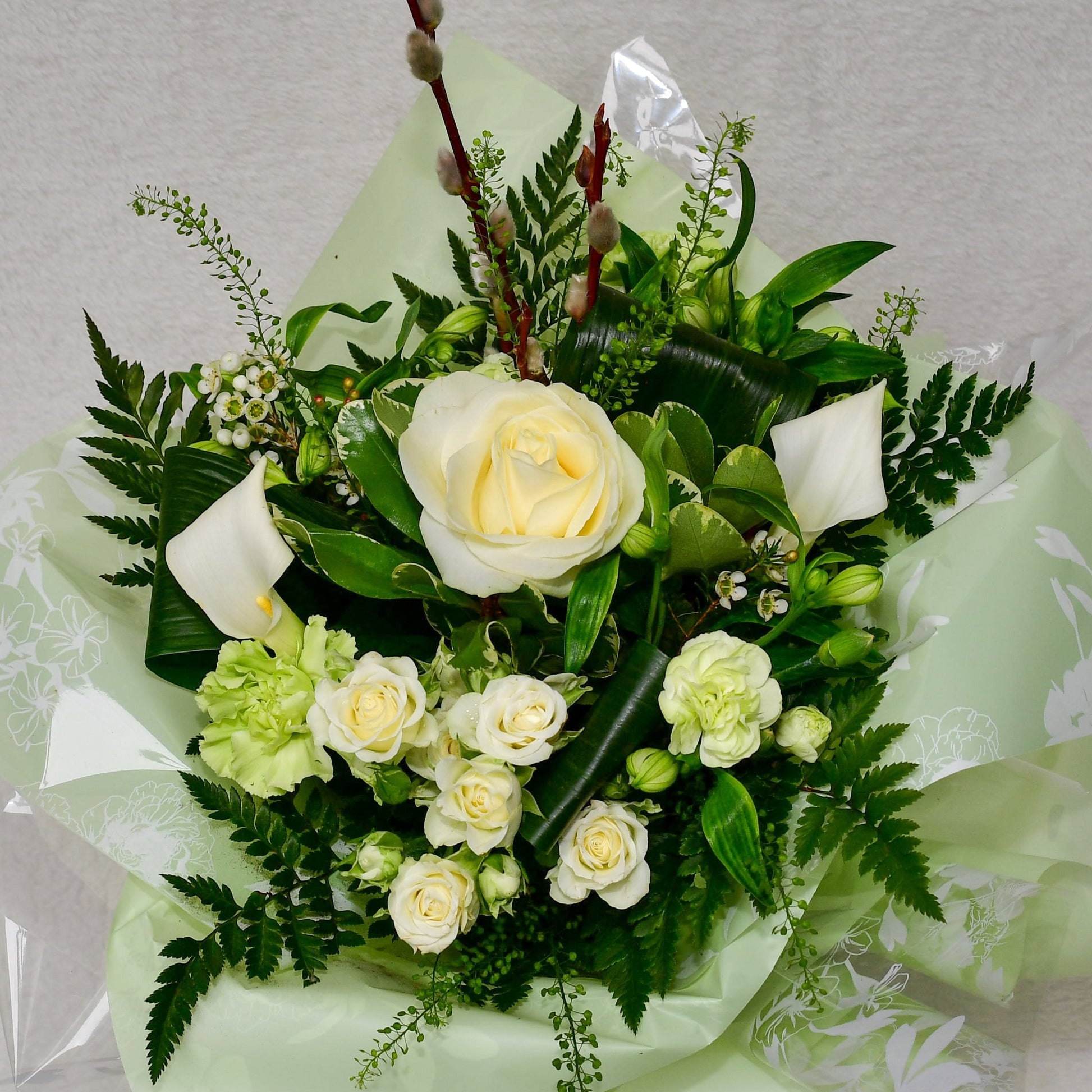 A floral bouquet with white and pale yellow roses, green hydrangeas, and green foliage, wrapped in pale green patterned paper and clear cellophane, photographed against a light gray textured background.