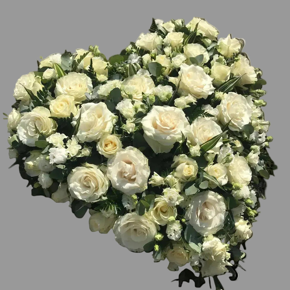 A heart-shaped floral arrangement of white and cream roses with green foliage, placed on a gray concrete surface.