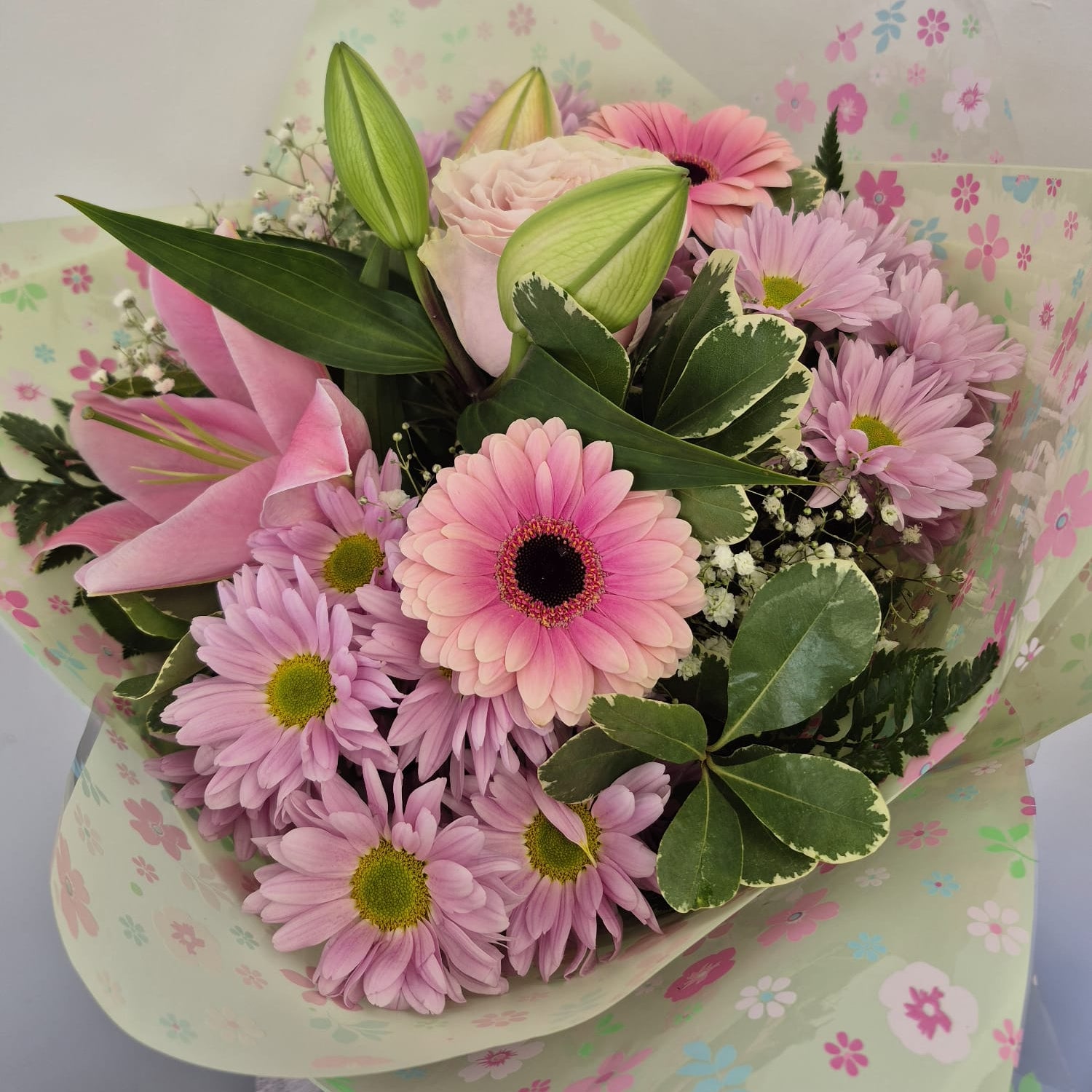A vibrant flower bouquet featuring pink gerbera daisies, pink lilies, a pale pink rose, and green foliage, wrapped in light green floral-patterned paper against a plain white background.