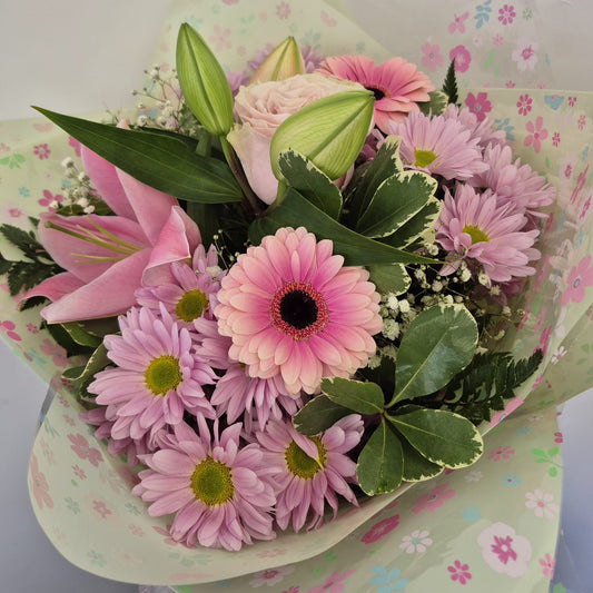 A vibrant flower bouquet featuring pink gerbera daisies, pink lilies, a pale pink rose, and green foliage, wrapped in light green floral-patterned paper against a plain white background.