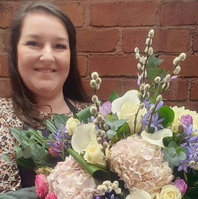 Katie holding a large, vibrant floral bouquet with pink, purple, cream, and white flowers, set against a red brick wall background.