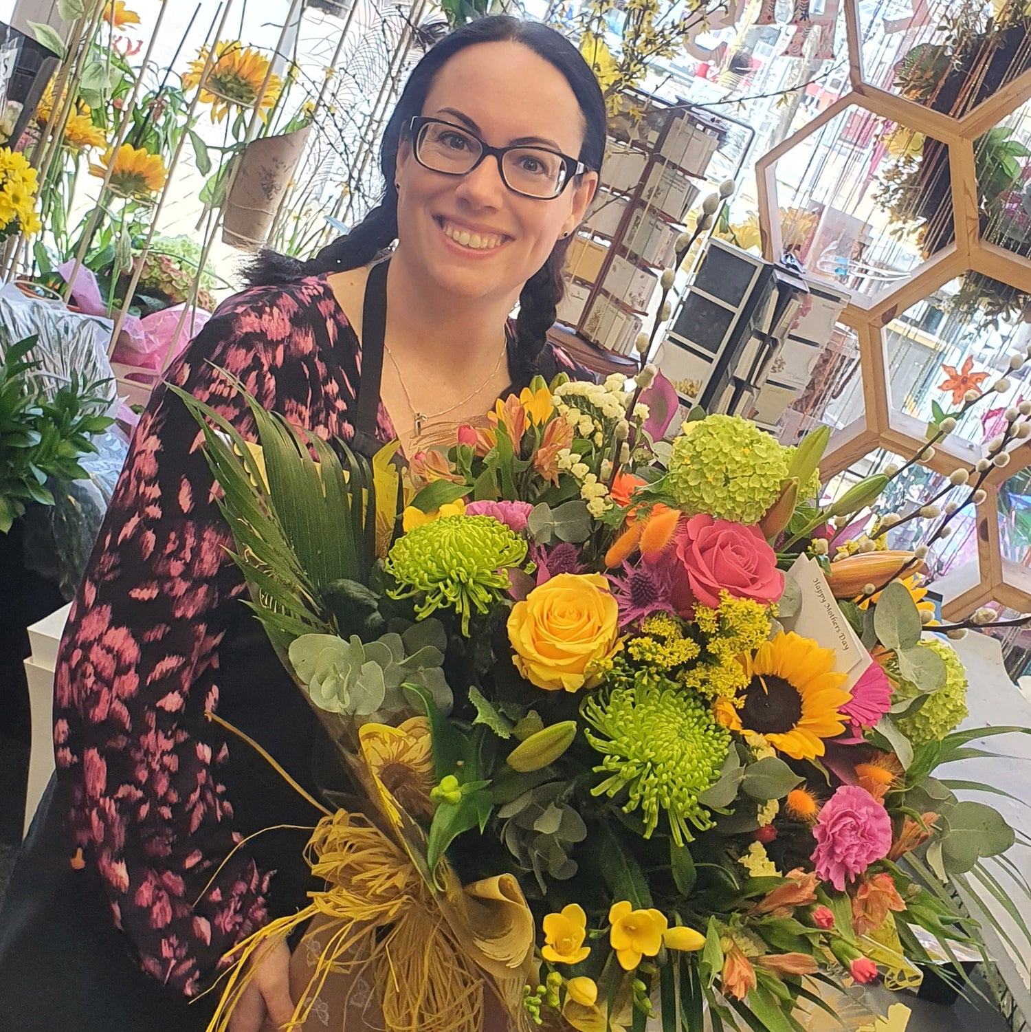 Laura Bacon holding a large, vibrant, multicolored flower bouquet in a flower shop. The bouquet features yellow roses, sunflowers, pink and orange blossoms, green chrysanthemums, and is wrapped in brown paper with a gold ribbon.