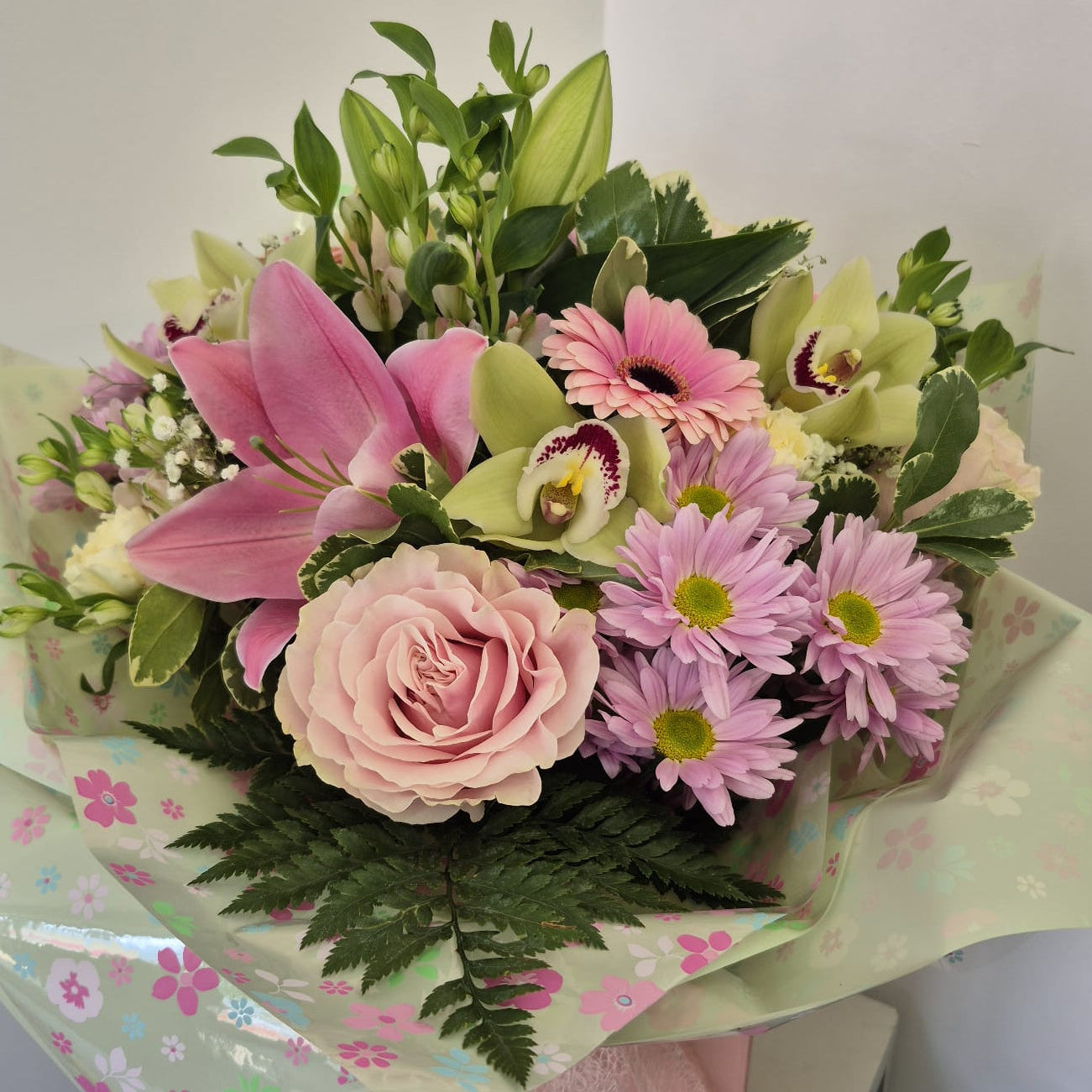A vibrant floral bouquet featuring pink lilies, a light pink rose, purple daisies, and green foliage, wrapped in a pale green floral-patterned paper and tied with a pink ribbon, resting on a light-colored surface against a plain wall.
